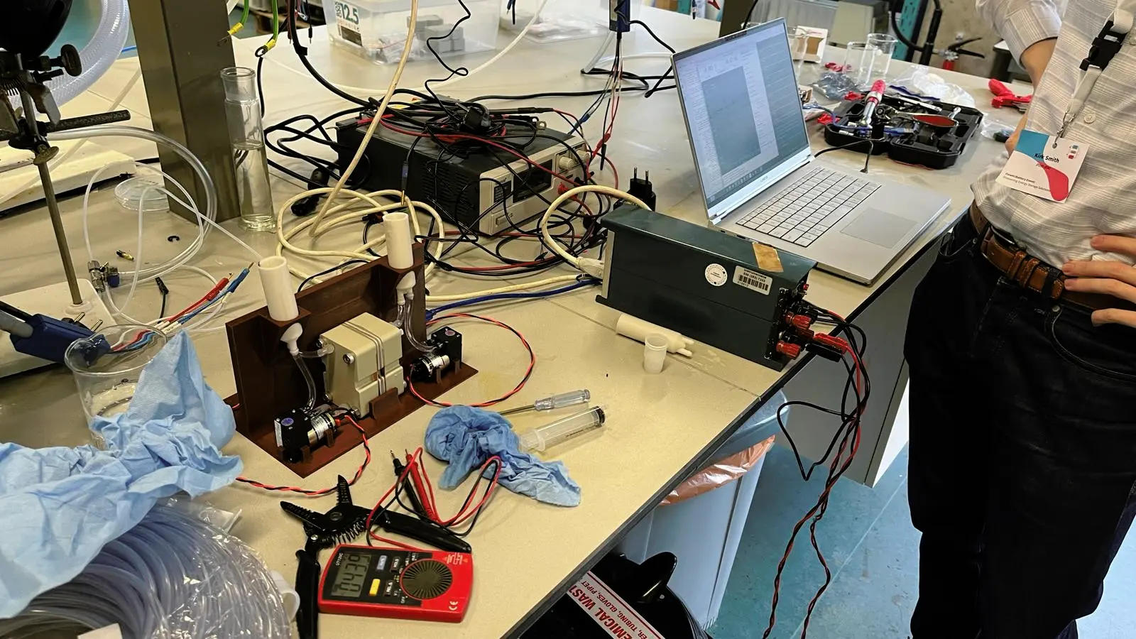 Flow battery test cell in jig on lab bench, with multimeter, power supply, and laptop nearby